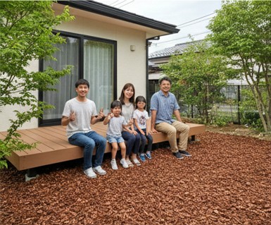 ウッドチップが敷き詰められた自然豊かな庭のウッドデッキで、笑顔でくつろぐ日本人の家族